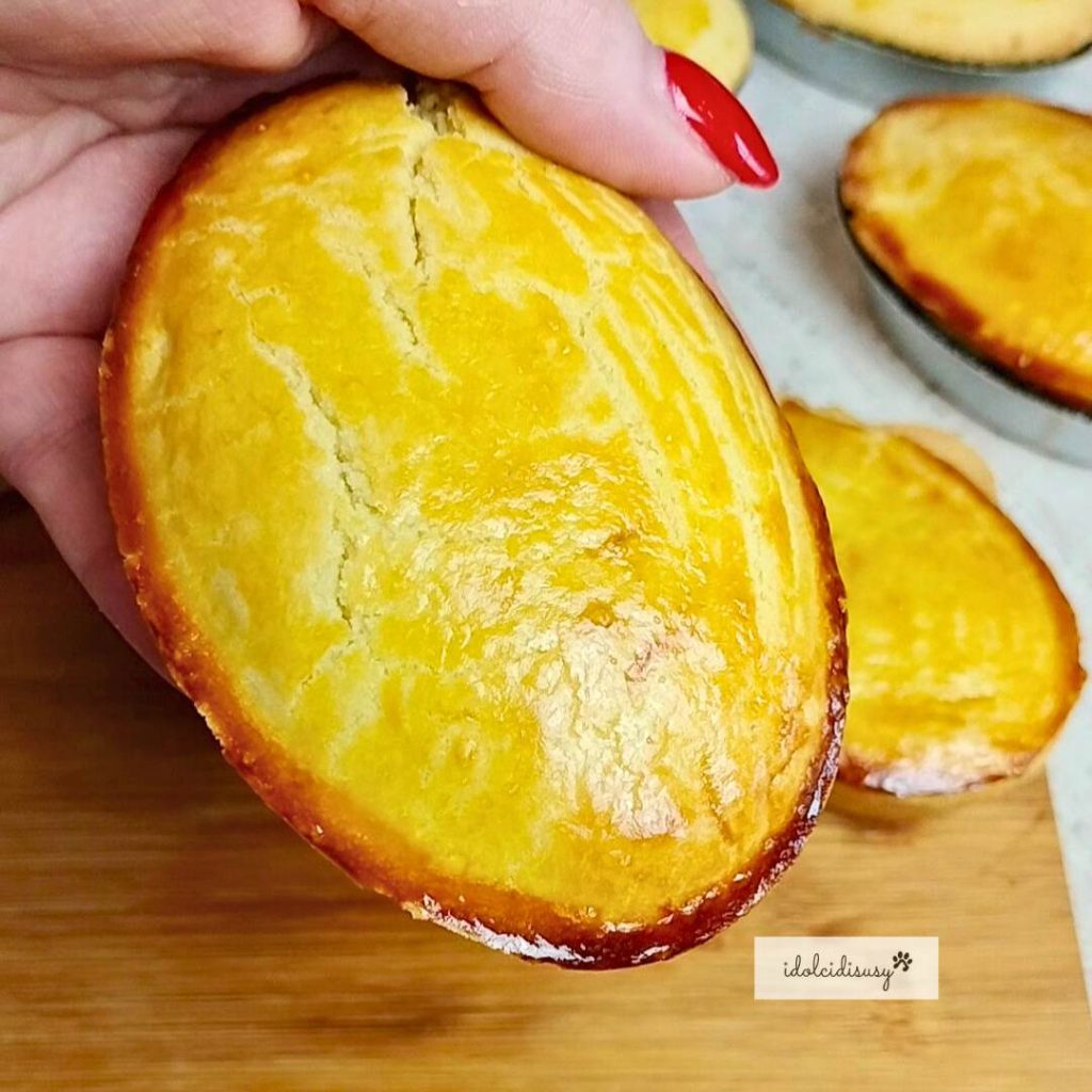 Whole oval pasticciotto with golden and shiny crust, just baked, held in hand and placed on a wooden cutting board.