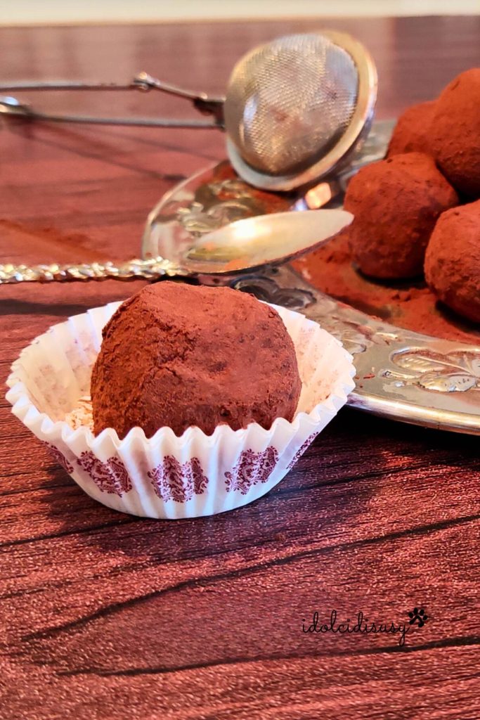 Close-up of a chestnut truffle covered in bitter cocoa in a decorated white cupcake liner, with plate and spoon in the background