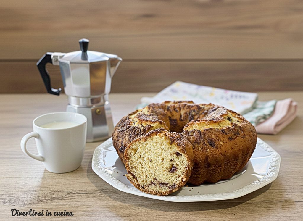 Tall and fluffy bundt cake with pieces of dark and milk chocolate, served on an elegant white plate. On the light wooden table, there is a metal moka coffee pot and a cup of white milk, creating a cozy and tidy breakfast atmosphere.
