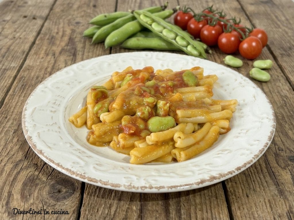 A plated dish of creamy casarecce with green fava bean cream and tomato sauce, garnished with fresh fava beans, on a rustic wooden table with fresh ingredients.