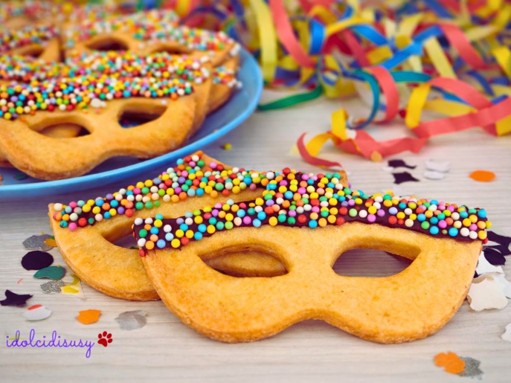 Karnevalskekse in Form einer Maske dekoriert mit Schokolade und buntem Zuckerstreusel, auf hellem Untergrund mit Konfetti