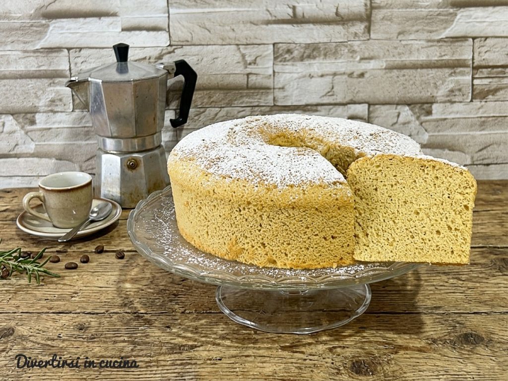 Eine lockere und hohe Chiffon Cake mit Kaffee auf einer eleganten Glas-Etagere, bestreut mit Puderzucker. Ein Stück wurde abgeschnitten und zeigt das luftige, weiche Innere des Kuchens, neben dem Hauptkuchen platziert.