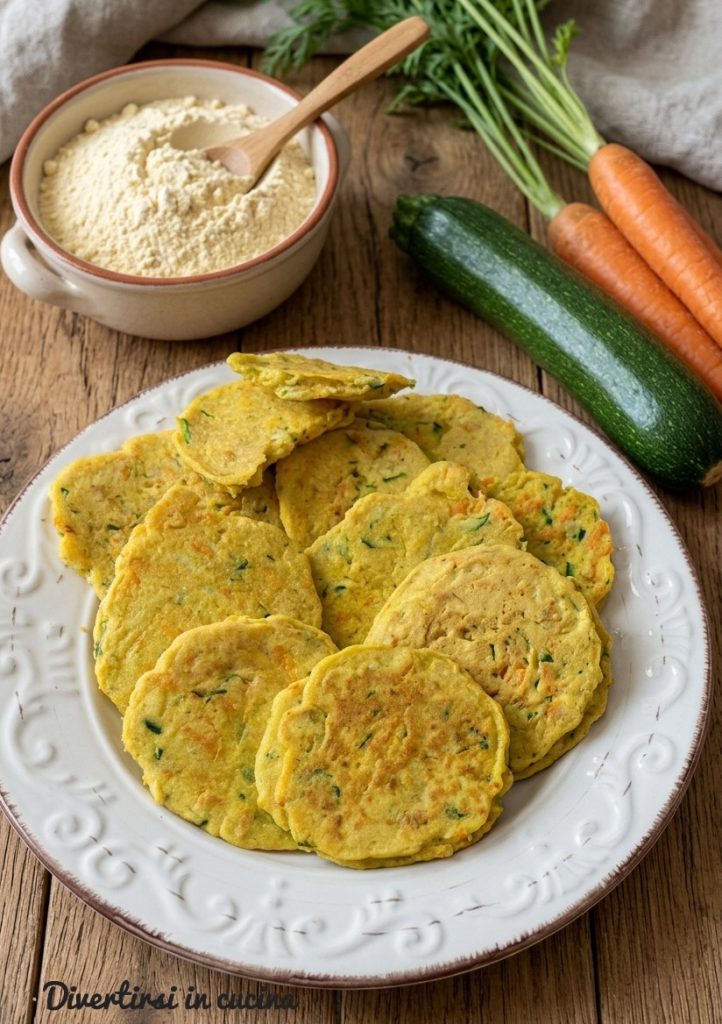Teller mit gelben Kichererbsen-Puffern mit geraspelten Zucchini und Karotten, neben frischen Zutaten auf einem Holztisch.