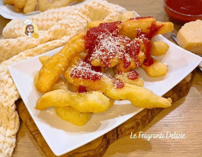 Teller mit neapolitanischen Engelchen, frittiert, goldbraun und knusprig, serviert auf einem rechteckigen weißen Teller, garniert mit Tomatensauce und einer großzügigen Prise geriebenen Käses. Im Hintergrund sind weitere Engelchen, eine Schälchen Sauce und ein Stück Käse auf einem Olivenholzbrett zu sehen. Rezept vom Blog Le Fragranti Delizie.