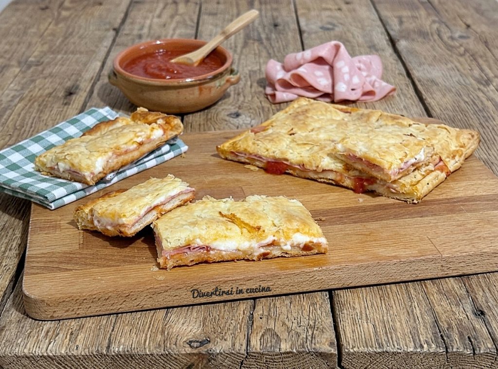 Ein Holzbrett mit aufgeschnittenen Stücken eines Rustico mit Blätterteig, Stracchino und Mortadella, daneben eine kleine Schale Tomatensauce und Scheiben Wurst auf einem rustikalen Holztisch