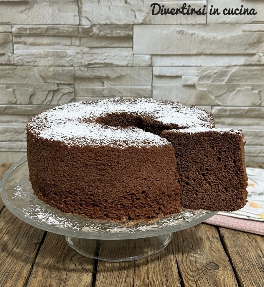 Une généreuse tranche de chiffon cake au cacao, découpée et placée à côté du gâteau entier, saupoudrée de sucre glace sur un présentoir en verre posé sur une table en bois rustique.