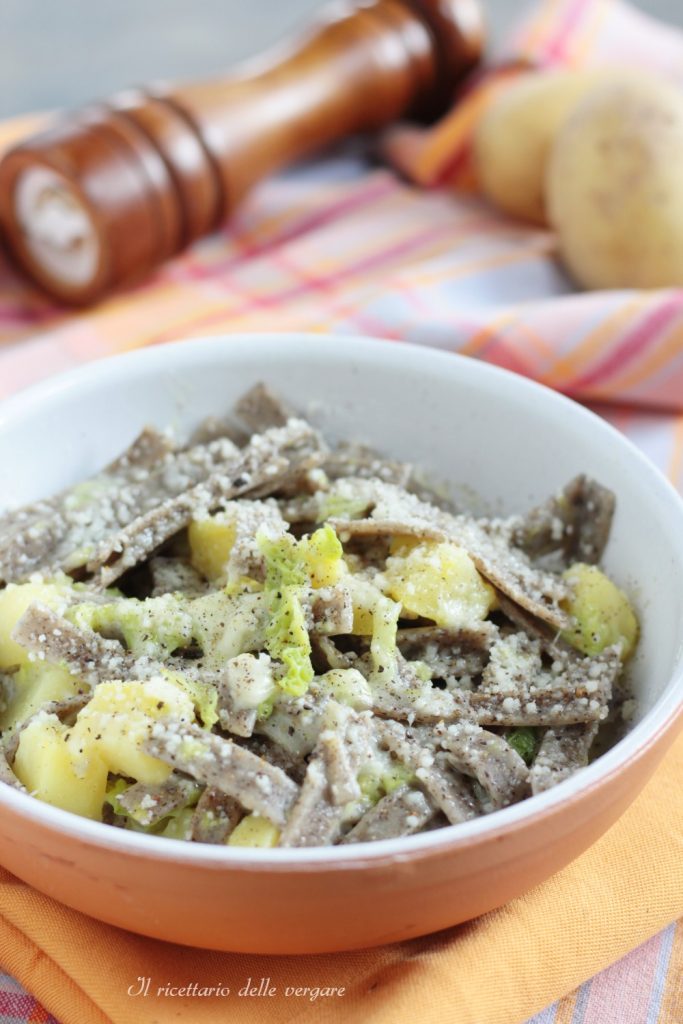 Plano vertical de una ración de pizzoccheri fundente con verza, patatas y fontina, servida en un plato rústico.