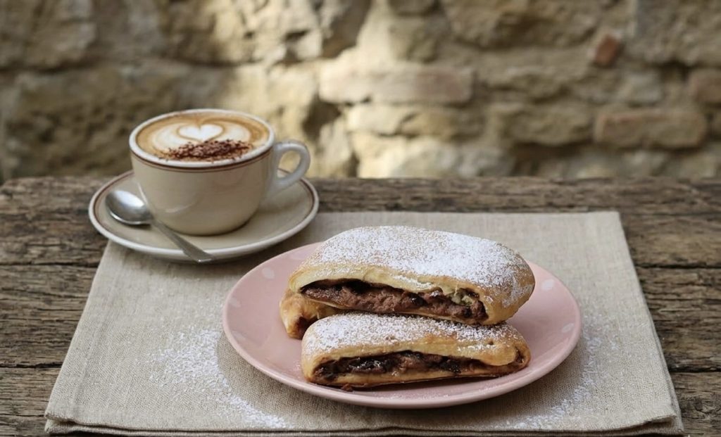 Dos saquitos de masa rellenos de chocolate sobre un plato rosa con lunares, colocado sobre un mantel de lino encima de una mesa de madera rústica. Al lado hay una taza de capuchino con latte art en forma de corazón, delante de una pared de piedra antigua