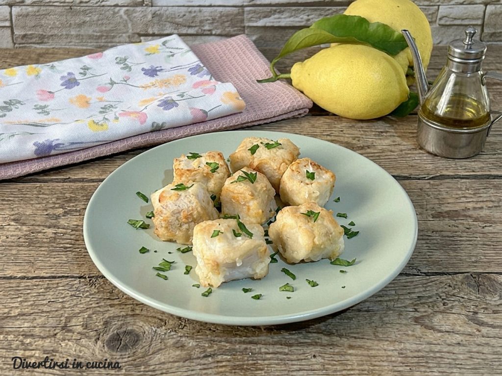 Um prato de cerâmica verde água com bocadinhos de merluza dourados e crocantes, guarnecidos com salsinha fresca, ao lado de limões inteiros e uma garrafinha de azeite sobre uma mesa de madeira