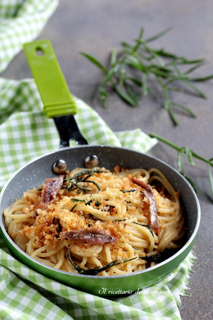 Plano vertical de espaguete com paccasassi e anchovas servidos em uma panelinha verde com ramos de funcho marinho ao fundo.