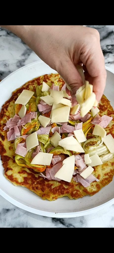 We draaien de pizza om en maken hem af met courgettebloemen, ham en scamorza
