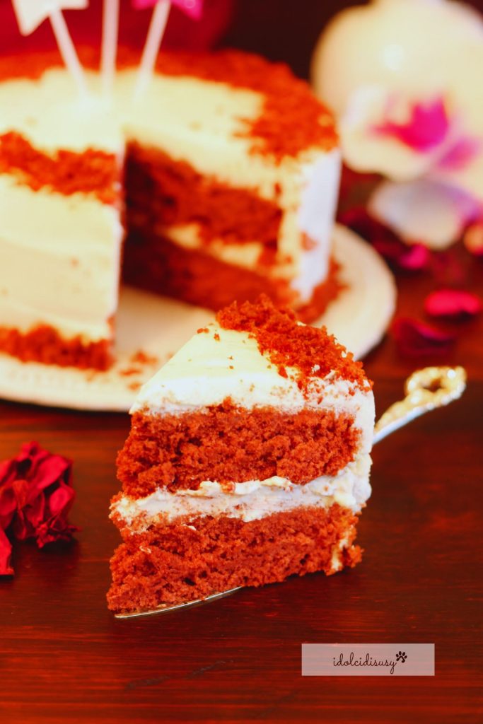 Een plak zelfgemaakte Red Velvet Cake, zacht en vochtig, met lagen rode biscuit en roomkaas, geserveerd op een schotel met de taart op de achtergrond